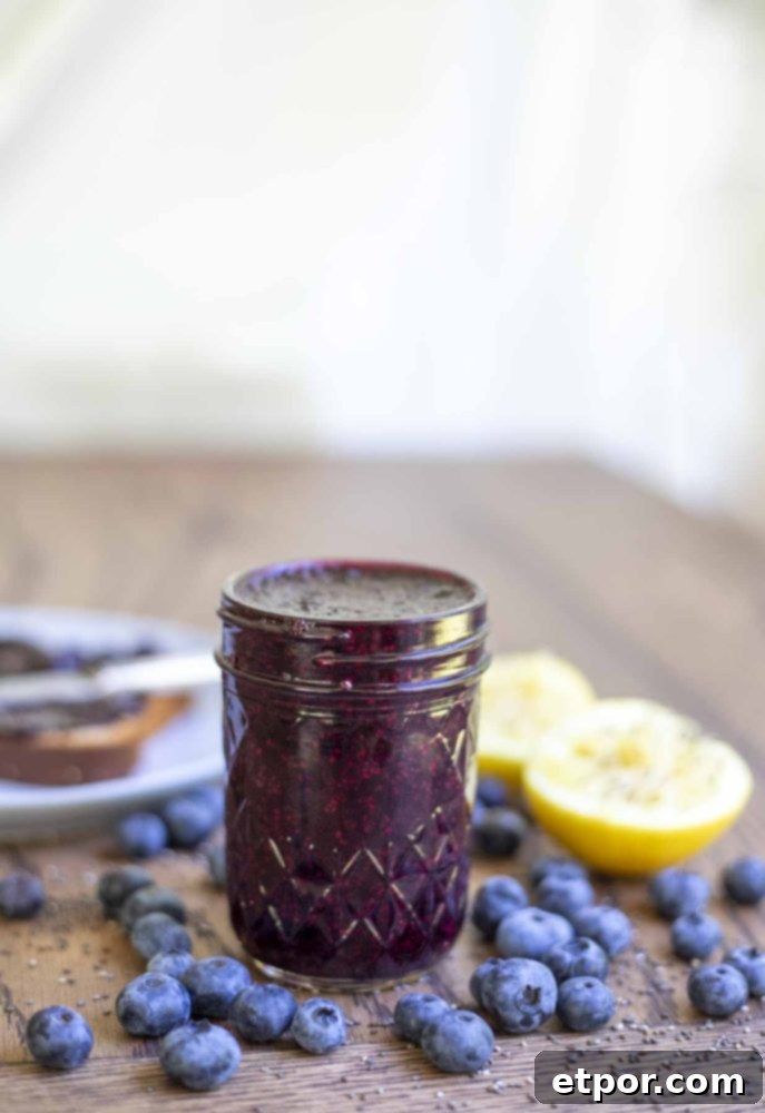 jar of blueberry freezer jam on a wood table surrounded by blueberries, sliced lemons and a plate with toast