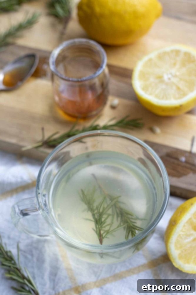 rosemary tea with fresh sprigs of rosemary in a glass mug with a cutting board with a jar of honey, lemon slices, and rosemary in the background