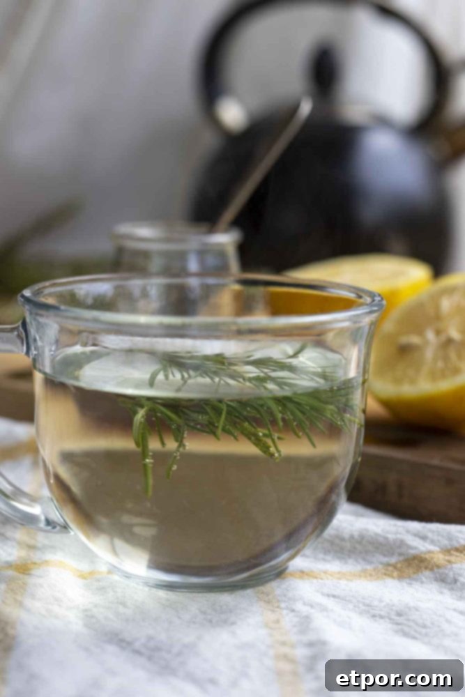 side view of a glass mug of rosemary tea on a white and yellow plaid towel with a cutting board of lemons and jars in the background