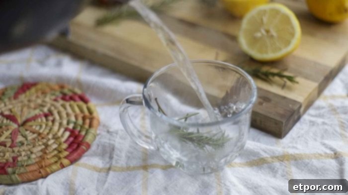 Pouring boiling water into a glass mug with rosemary sprigs. The mug sits on a yellow and white towel with a cutting board in the background