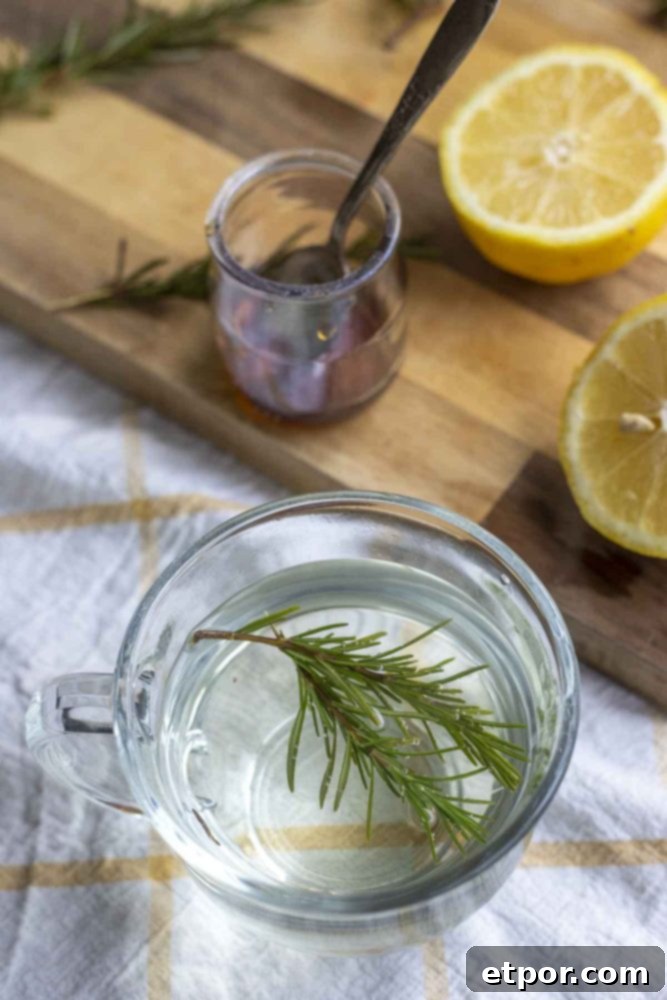 fresh sprigs of rosemary in a glass mug on a white and yellow towel with a wood cutting board with sliced lemons and a jar of honey in the background