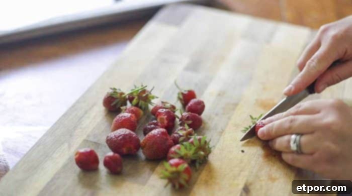 A hand using a paring knife to hull and slice fresh strawberries on a wooden cutting board.