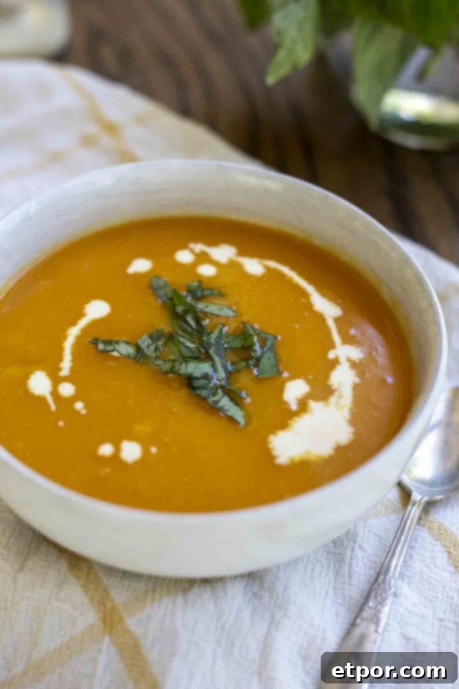 A beautifully presented bowl of 3-ingredient butternut squash soup, drizzled with cream and garnished with fresh basil. The white bowl sits on a white and yellow checkered towel, inviting a warm and comforting meal.