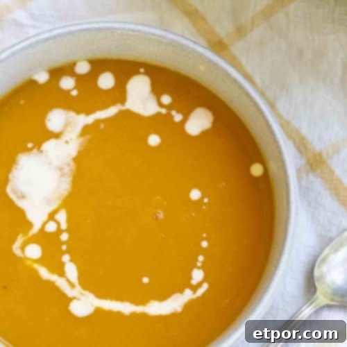 An overhead photo showing a bowl of 3-ingredient butternut squash soup, elegantly drizzled with cream. The bowl rests on a white and yellow checkered towel, with a spoon positioned invitingly to the right.