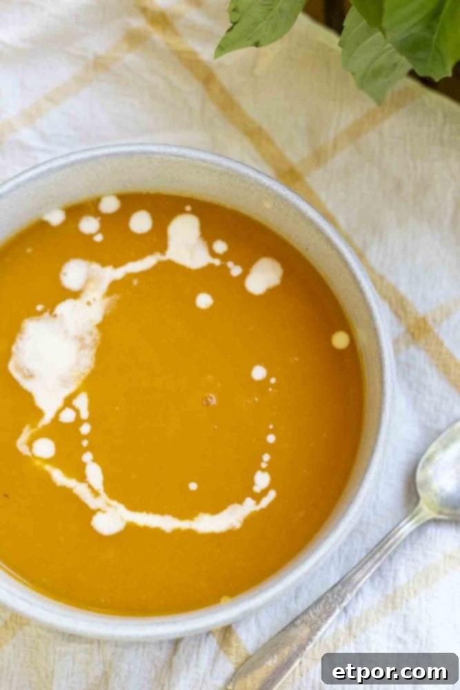 An overhead shot of a bowl of 3-ingredient butternut squash soup, adorned with a delicate swirl of cream, resting on a white and yellow checkered towel. A spoon lies to the right, inviting a taste.