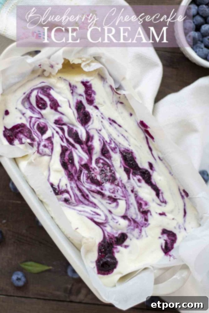 Velvety No-Churn Blueberry Cheesecake Ice Cream 2 Overhead photo of a loaf pan lined with parchment paper, filled with creamy blueberry cheesecake ice cream, showcasing beautiful swirls of vibrant blueberry sauce. The pan rests on a white towel, surrounded by scattered fresh blueberries.