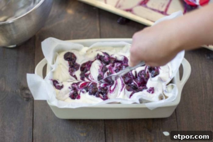 Velvety No-Churn Blueberry Cheesecake Ice Cream 5 A hand gently swirls blueberry sauce into creamy cheesecake ice cream using a knife. The ice cream is set in a loaf pan, lined with parchment paper, on a warm wooden table.