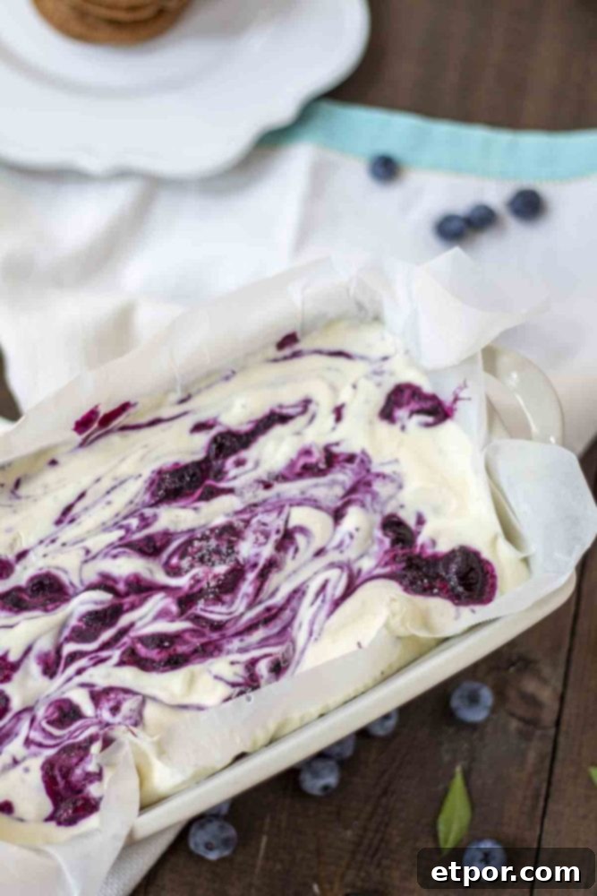 Velvety No-Churn Blueberry Cheesecake Ice Cream 6 A close-up of blueberry cheesecake ice cream in a loaf pan, resting on a white and teal striped towel. Fresh blueberries are charmingly scattered around the pan on the wooden table, highlighting the vibrant fruit.