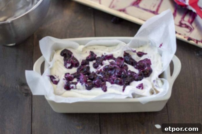 Velvety No-Churn Blueberry Cheesecake Ice Cream 8 Luscious blueberry sauce dolloped over the creamy cheesecake ice cream base in a parchment-lined loaf pan, waiting to be swirled. This beautiful layer rests on a wooden table.
