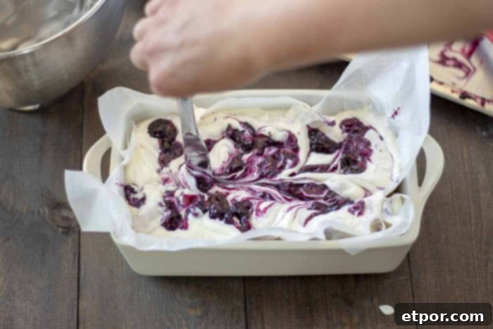 Velvety No-Churn Blueberry Cheesecake Ice Cream 9 A hand holds a knife, artfully creating zig-zag swirls in the blueberry cheesecake ice cream mixture within a parchment-lined loaf pan. The pan is situated on a charming wooden table, ready for freezing.