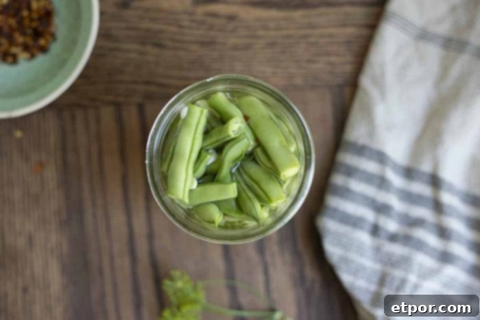 overhead photo of an open jar of quick pickled green beans on a wooden table with a blue and white stripped towel to the right, and a teal bowl of crushed red peppers to the left
