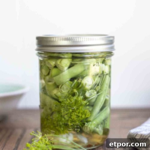 pickled green beans in a mason jar with a lid on a wood table. Fresh dill sits in front of the jar.