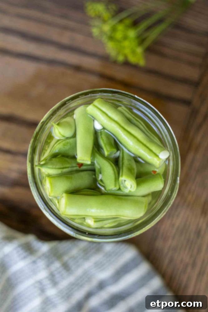 overhead photo of pickled green beans in a mason jar on a wood table with a blue and white towel in front