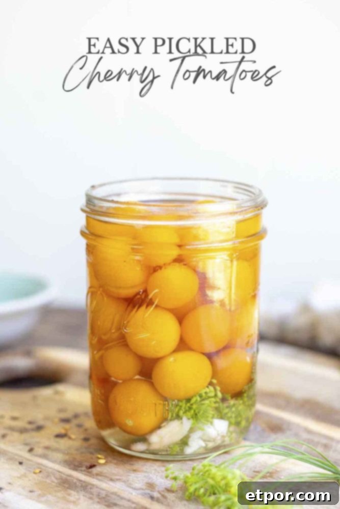 pint mason jar full of pickled cherry tomatoes with garlic and dill in a brine. The jar sits on a wood countertop with red pepper flakes and dill