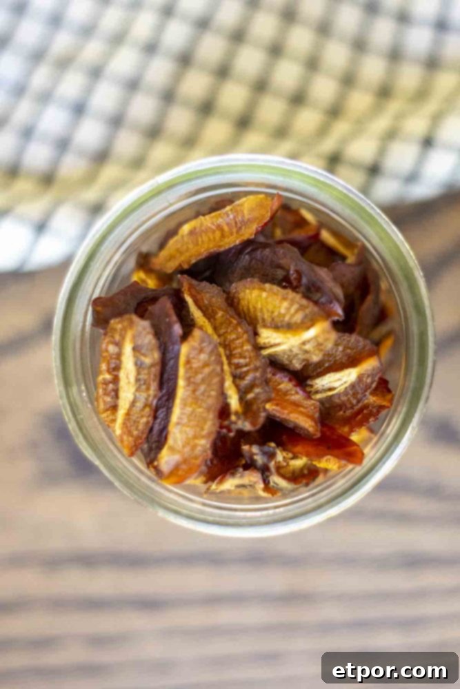 Overhead photo of dehydrated plums in a glass jar on a wood countertop with a white and black checked towel in the background.
