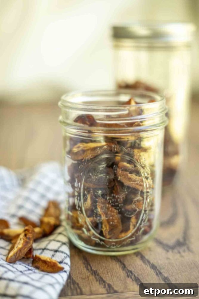 Jar of dried plums on a wood table with another jar in the background. A black and white towel with more prunes is to the right.