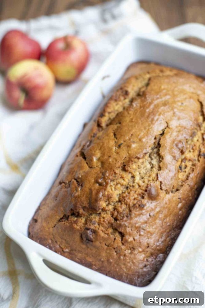 Freshly baked sourdough apple bread still in its loaf pan, resting on a wooden cutting board with a yellow and white towel and whole apples in the background.