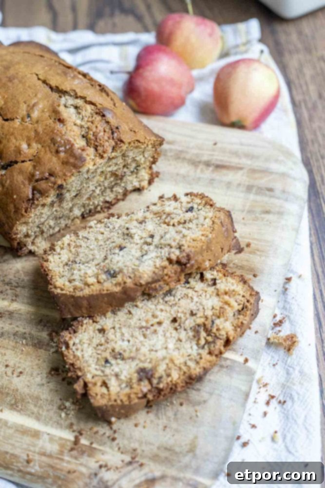 A loaf of sourdough apple bread with two slices cut off on a wood cutting board, placed on a white towel with fresh apples in the background.