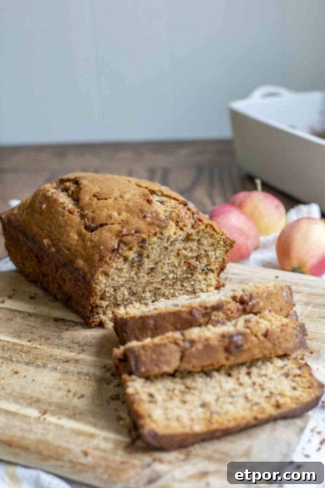 Loaf of sourdough cinnamon apple bread sliced on a wood cutting board with whole apples and a loaf pan in the background.