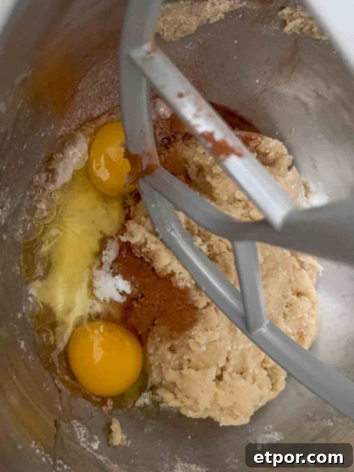 Eggs, cinnamon, baking soda, and baking powder being added to the fermented sourdough dough in a stand mixer bowl with a paddle attachment.