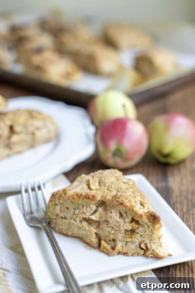 A single sourdough apple scone, beautifully glazed and resting on a white square plate, accompanied by a fork. In the soft background, another scone, a baking sheet, and fresh apples complete the inviting scene.