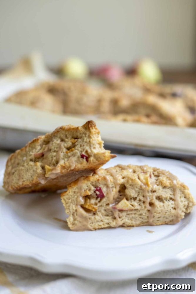 Two golden-brown sourdough apple scones stacked elegantly on a white plate, with a baking sheet filled with more delicious scones in soft focus behind them.