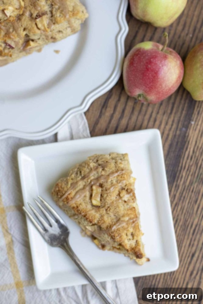 A single sourdough apple scone with a generous drizzle of cinnamon glaze, artfully placed on a white square plate with fresh apples and another scone in the background, creating a rustic and inviting display.