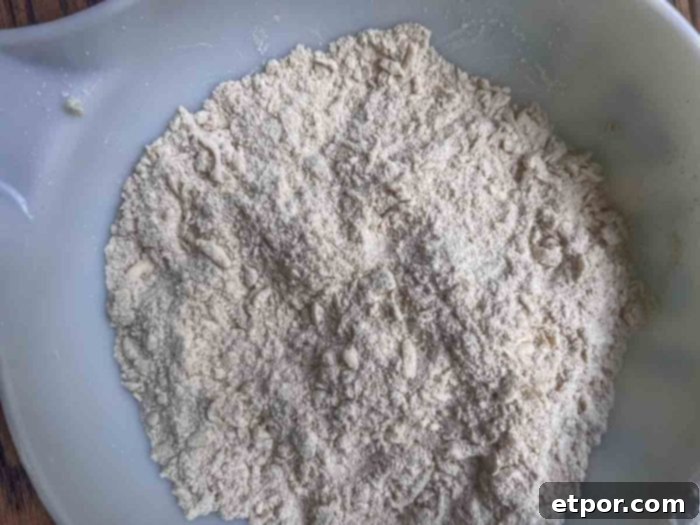 A white bowl holds a mixture of dry ingredients—flour, cinnamon, baking powder, and brown sugar—alongside small pieces of cold butter, ready to be combined.