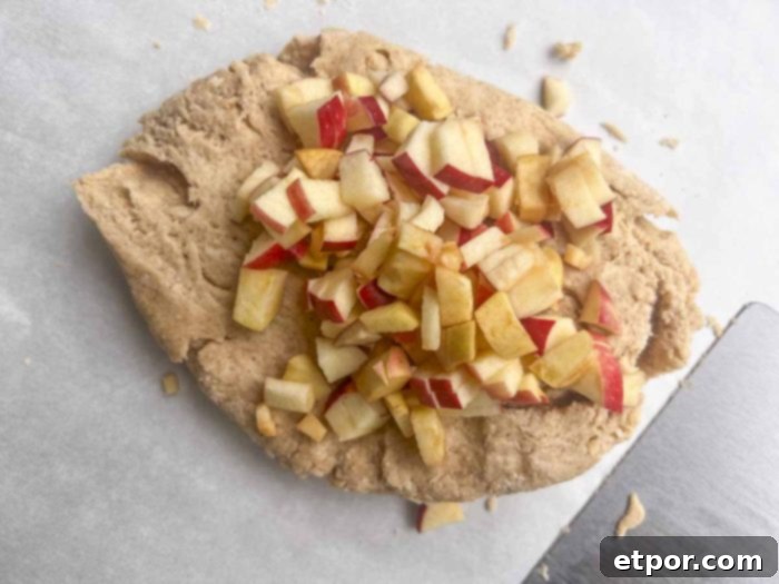Scone dough, spread on parchment paper, is generously topped with finely diced apples, ready to be kneaded and shaped for baking.