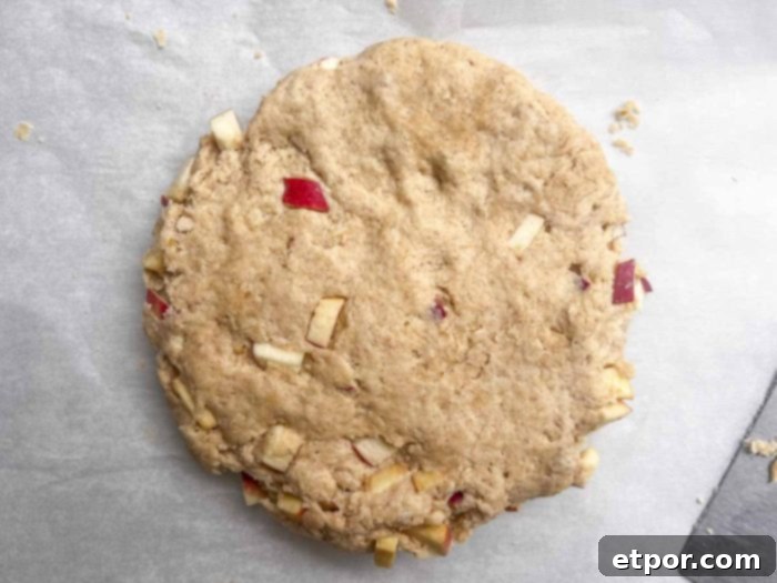 A circular disk of sourdough scone dough, studded with apple pieces, expertly cut into eight even wedges on parchment paper, awaiting the oven.