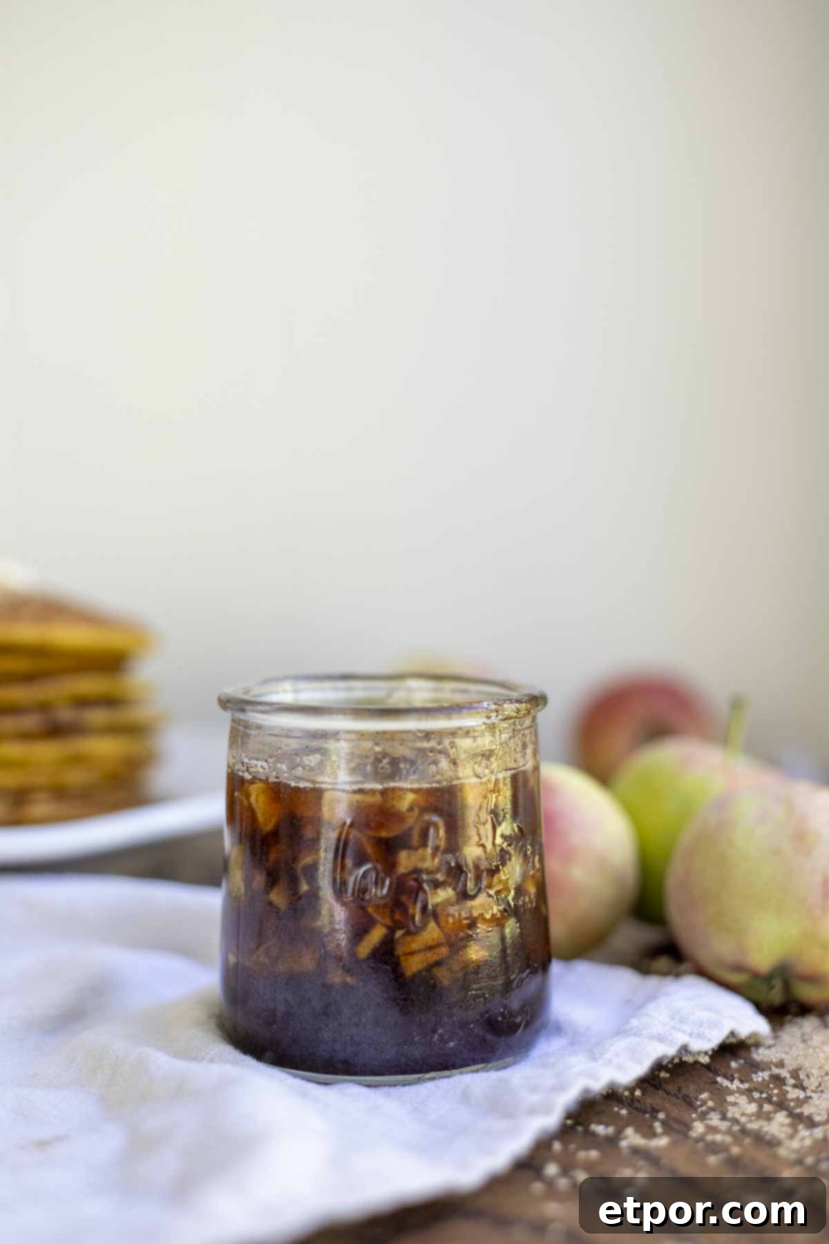 cinnamon apple syrup in a small glass jar on a napkin with apples and a stack of pumpkin pancakes in the background.