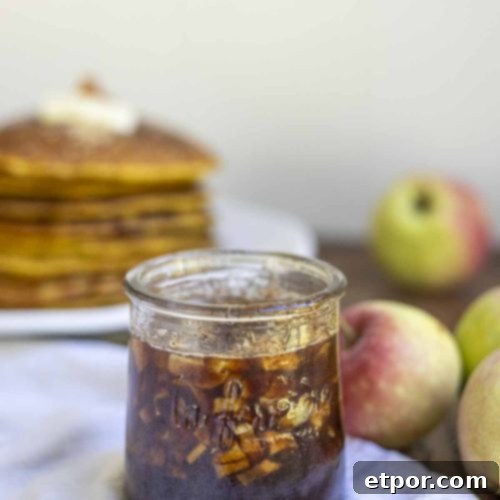 cinnamon apple syrup in a small glass jar on a napkin with apples and a stack of pumpkin pancakes in the background