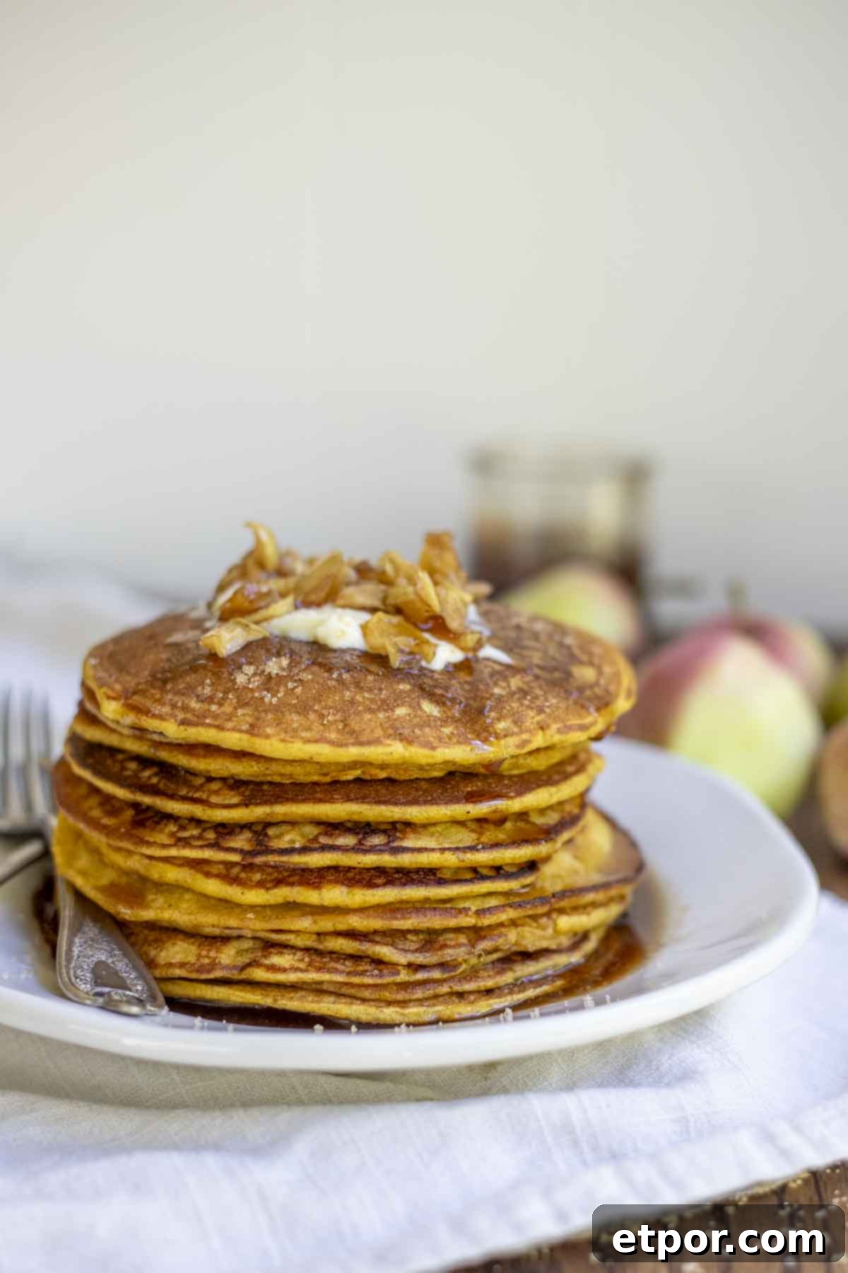 close up picture of a stack of sourdough pumpkin pancakes topped with butter, apples, and syrup on a white plate on a tan napkin.