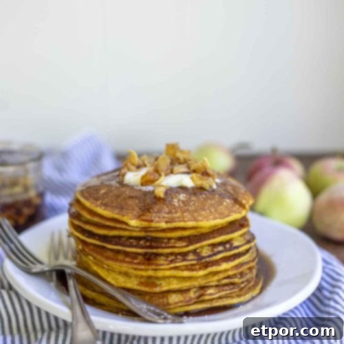 a stack of sourdough pumpkin pie pancakes topped with butter and apples on a white plate. The plate is on a white and blue stripped towel