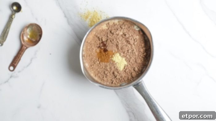 milk, spices, cocoa powder, and maple syrup in a small saucepan on a marble counter with measuring spoons in the top left corner.