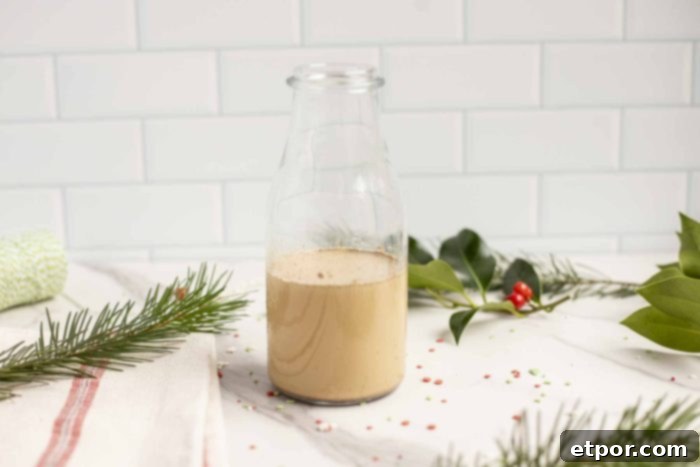 Whip Up Homemade Gingerbread Coffee Creamer 3 gingerbread coffee creamer in a glass milk jar on a marble countertop surrounded by greenery and holly.