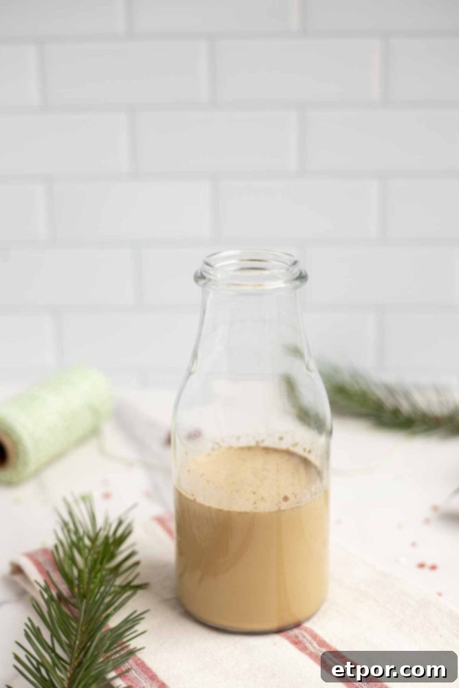 Whip Up Homemade Gingerbread Coffee Creamer 10 a small milk jar of gingerbread creamer on a white and red stripped towel with greenery to the left and a spoon of green thread in the background