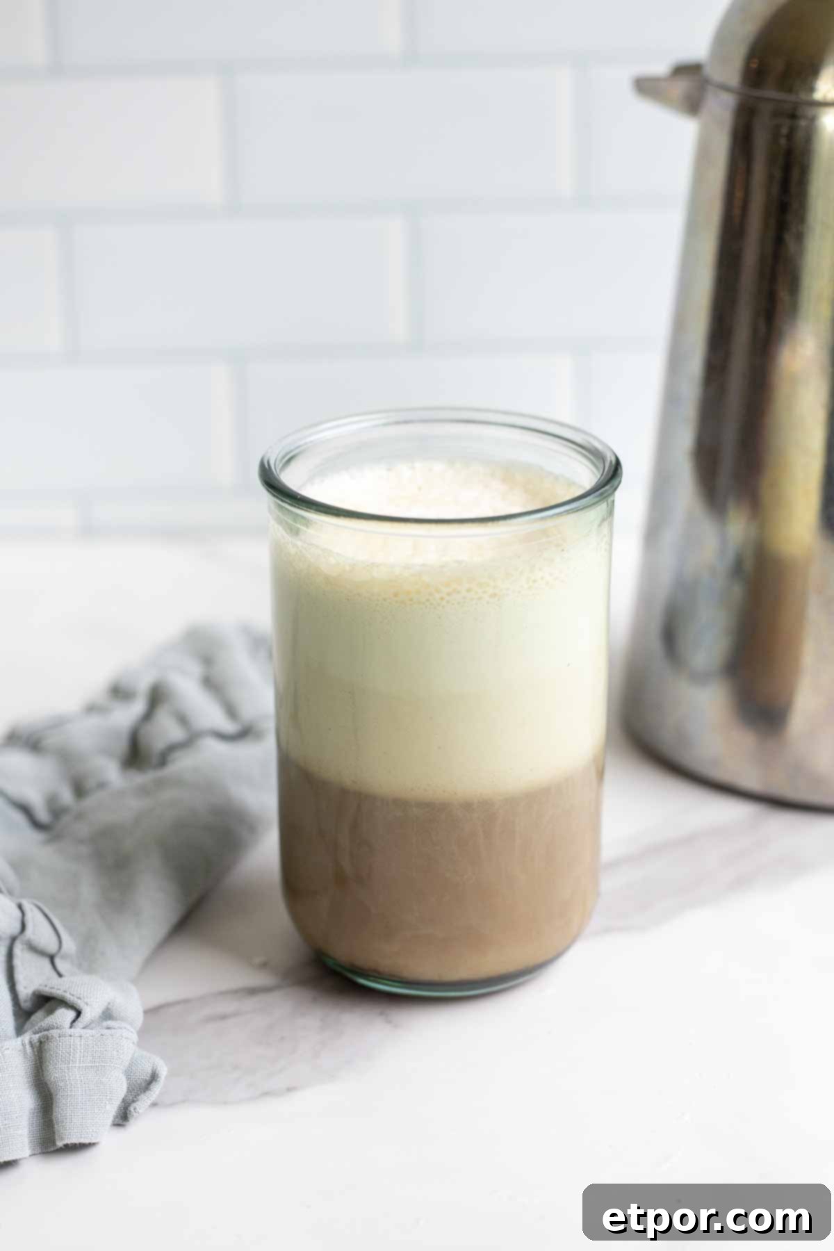 a glass of coffee topped with sweet cream cold foam on a marble countertop with a blue napkin and a metal French press in the background.