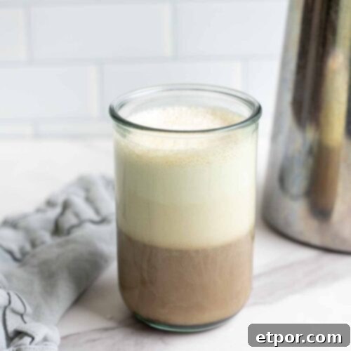a glass of coffee topped with sweet cream cold foam on a marble countertop with a blue napkin and a metal French press in the background.