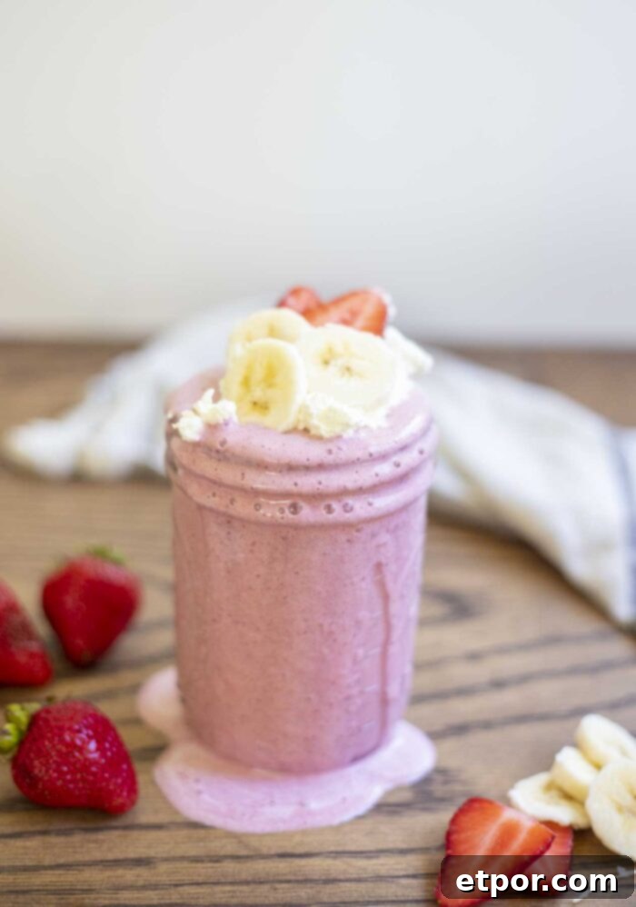 A vibrant strawberry banana milkshake in a mason jar, with creamy drips on the sides. It's garnished with fresh whipped cream, sliced strawberries, and banana slices, surrounded by more fresh fruit and a soft towel in the background.