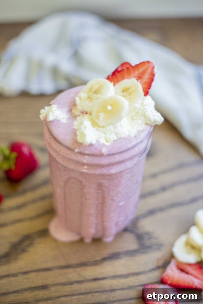 Close-up of a strawberry banana milkshake in a mason jar, artfully topped with fluffy whipped cream, fresh banana slices, and ruby-red strawberry slices. The jar rests on a rustic wood table, adorned with whole and sliced strawberries and a towel in the background.