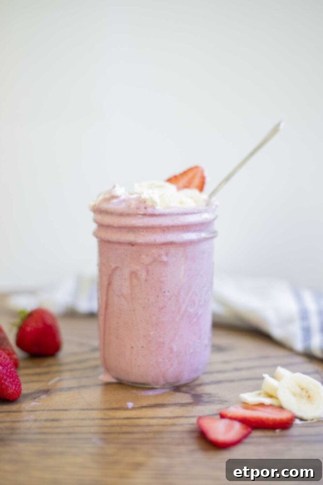 A healthy strawberry banana milkshake in a mason jar, garnished with whipped cream, banana slices, and strawberry slices. The jar sits on a warm wood countertop, surrounded by neatly arranged whole and sliced strawberries and banana slices.