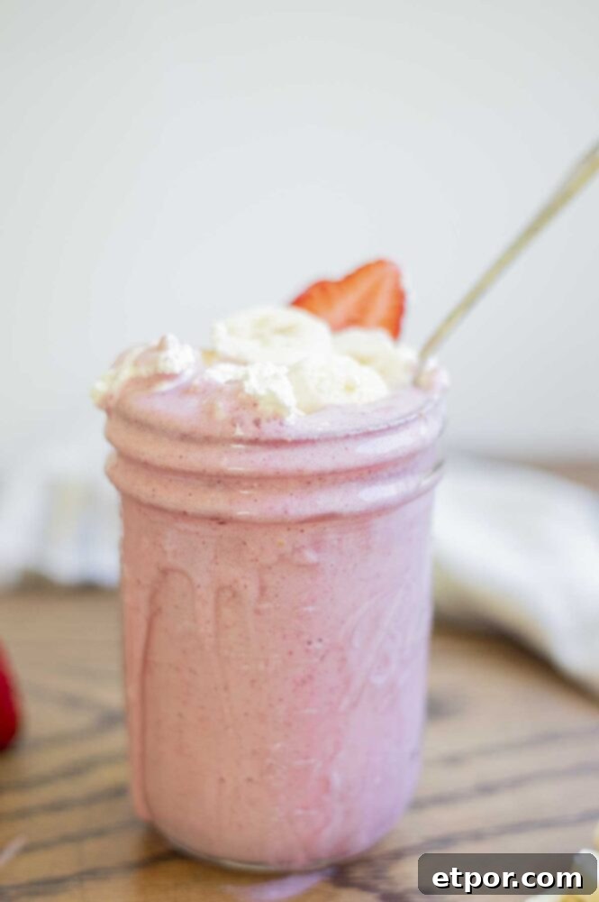 A healthy strawberry banana milkshake in a mason jar with a spoon, generously topped with whipped cream, fresh banana slices, and strawberry slices. A soft, cream-colored towel is visible in the background, adding to the cozy aesthetic.