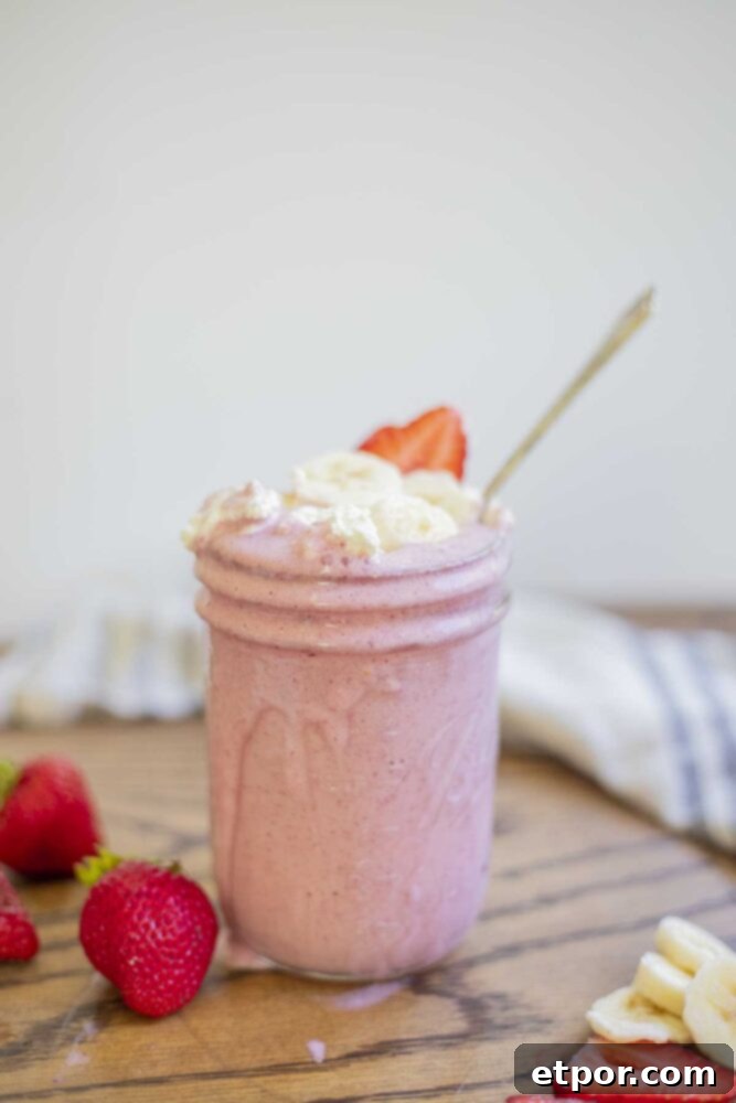 A beautiful strawberry banana milkshake in a mason jar with delightful drips along the sides. It's elegantly topped with fresh whipped cream, perfectly sliced strawberries, and banana. Surrounding the jar are an assortment of fresh strawberries and bananas, with a cozy towel in the background.