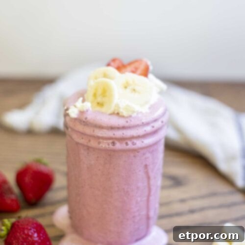 A vibrant strawberry banana milkshake in a mason jar, with creamy drips on the sides. It's garnished with fresh whipped cream, sliced strawberries, and banana slices, surrounded by more fresh fruit and a soft towel in the background.