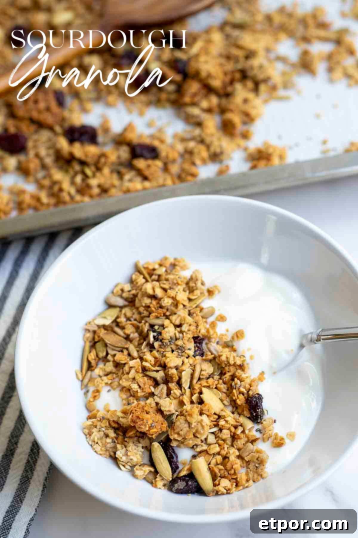 A white bowl of yogurt topped with sourdough granola. A baking sheet of more granola is in the background.