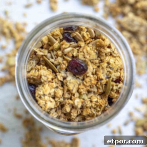 overhead photo of sourdough granola with seeds and dried cranberries in a mason jar. The jar is on a parchment lined baking sheet with more granola