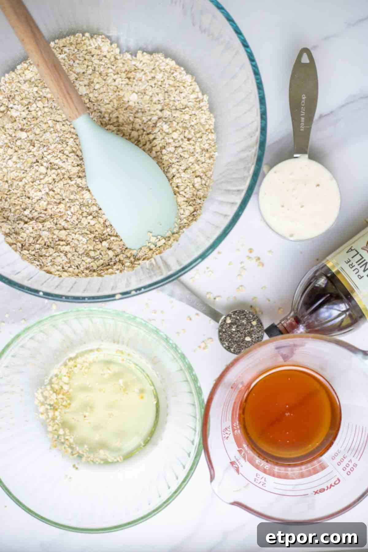 Glass bowls of oats, oil, honey, and measuring cups of sourdough starter and chia seeds and a bottle of vanilla extract are on a marble countertop.