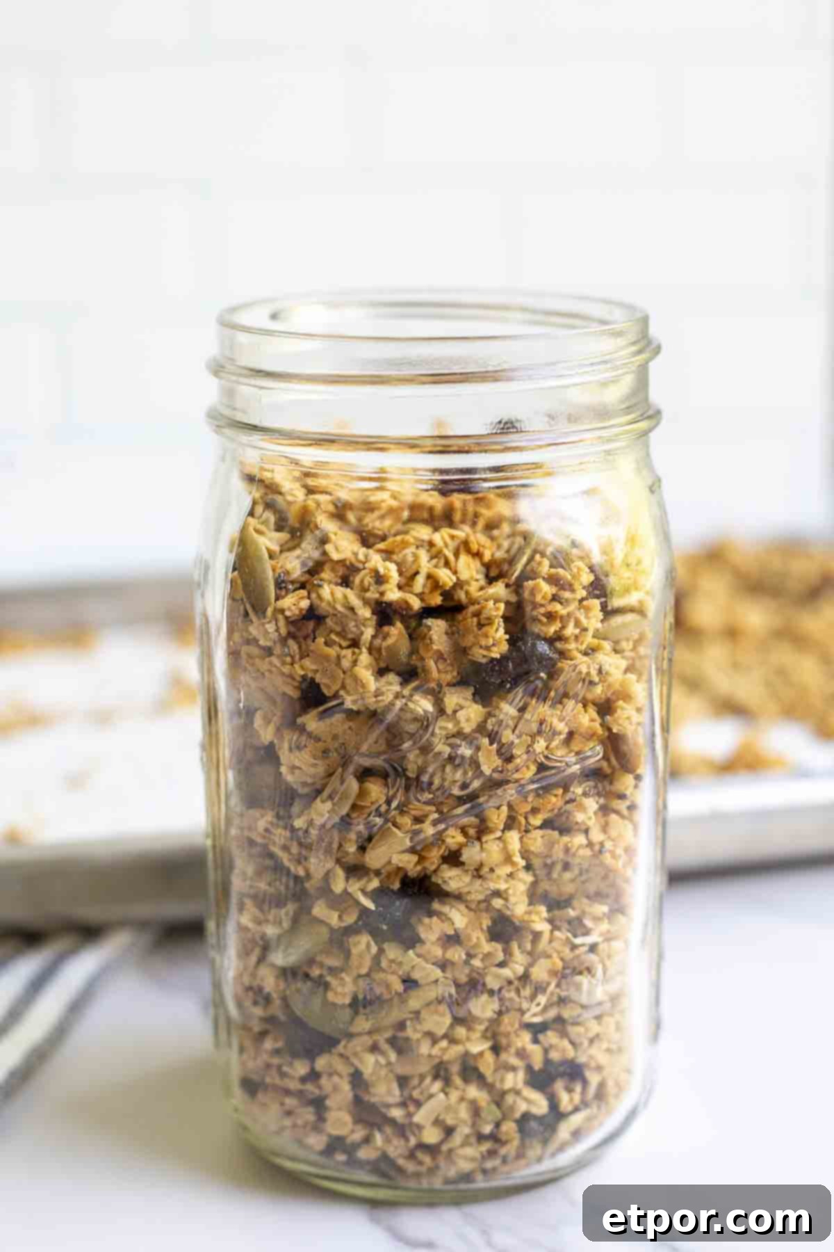 A mason jar of sourdough granola on a marble counter with a baking sheet of more granola in the background.