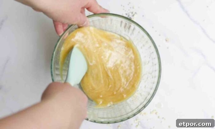 Sourdough starter, honey, and oil in a glass bowl getting mixed together with a silicon spatula.